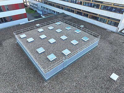 Drohnen-Blick von oben auf das kiesbedeckte Dach der Hochschul-Bibliothek in Rüsselsheim. Auf der erhöhten quadratischen, mit einem Geländer gesicherten Ebene stehen 16 Flachdachfenster essersky®  hevor, deren Glas bläulich schimmert.  